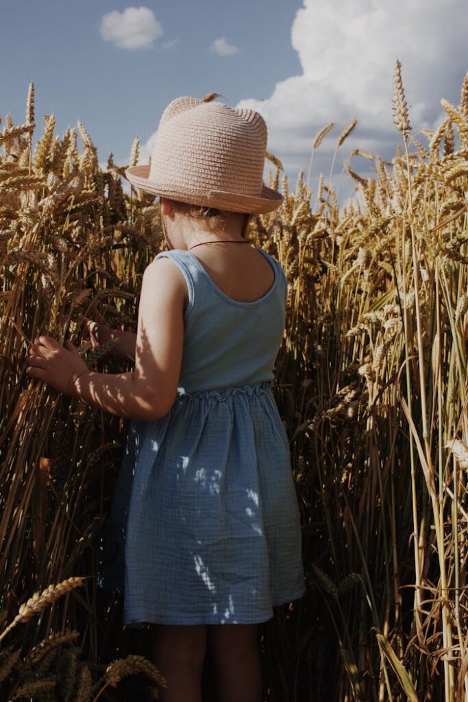 A young girl in a blue dress and straw hat stands in a golden wheat field.